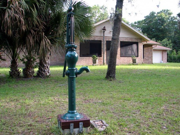 Back view of house. Working hand pump, farm bell on post at back door.